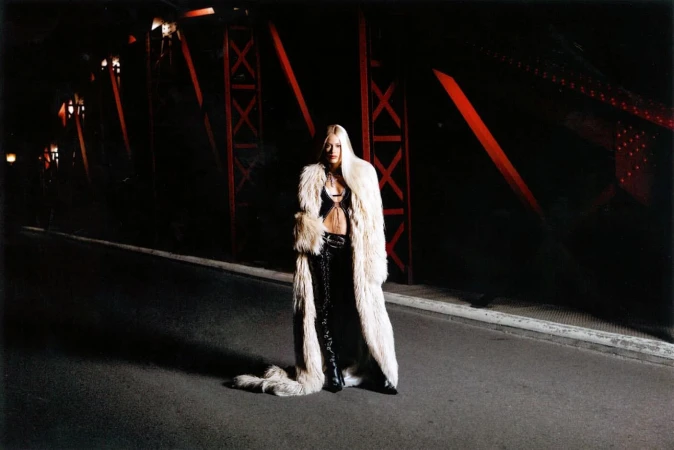 Adult female in a fur coat standing on a bridge at night, illuminated by low lighting with steel beams in the background.