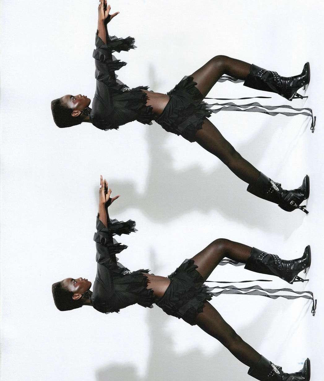 Fashion editorial shot of an African-American woman in a ruffled, dark outfit and high-heeled boots posing against a white background.