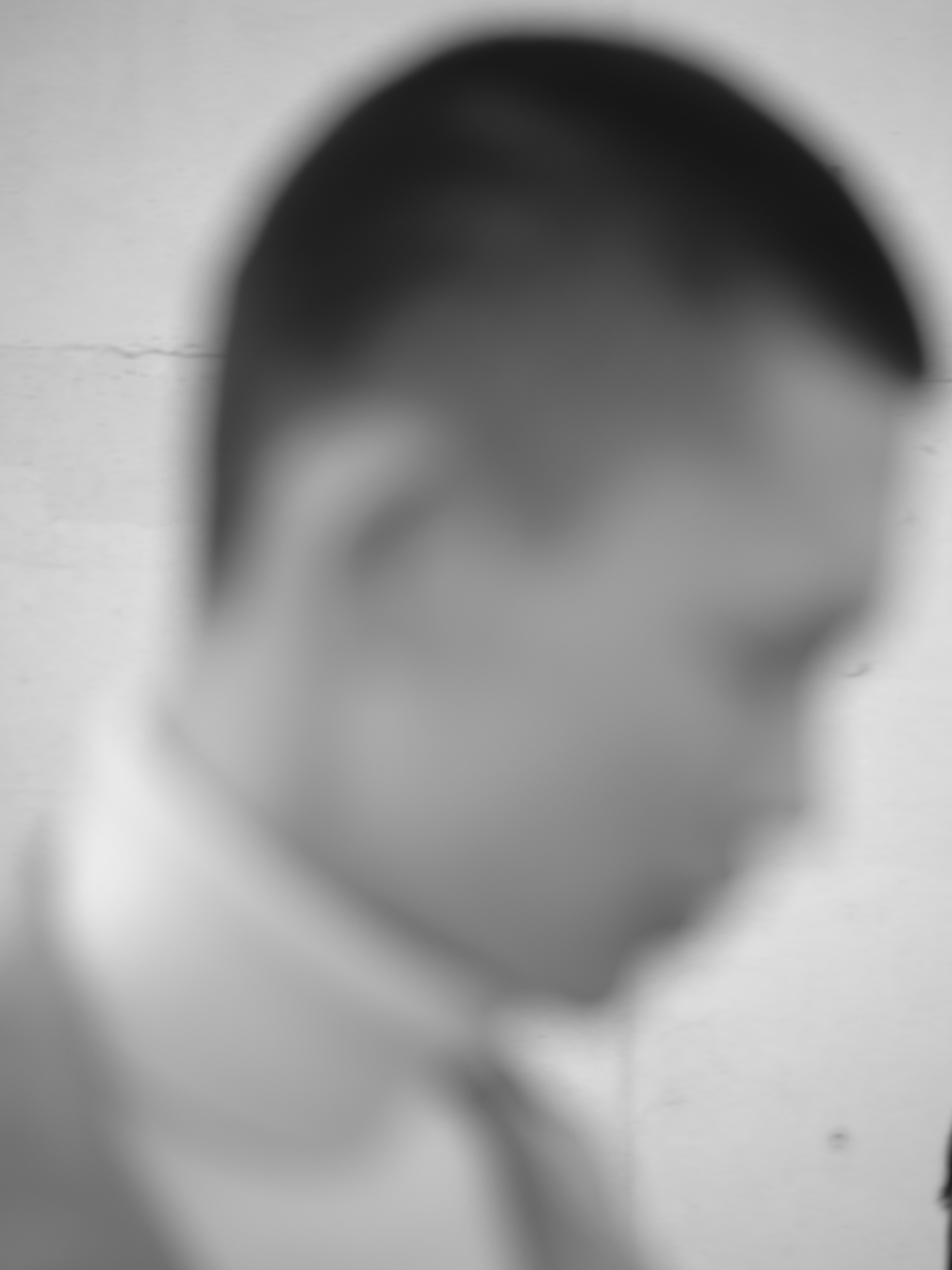 Blurry grayscale portrait of an adult male with short dark hair wearing a white collared shirt and jacket against a plain background.