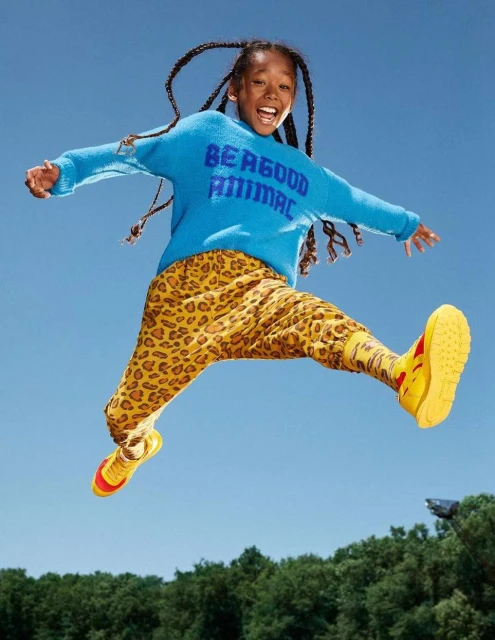 A happy child with braids jumps against a blue sky, wearing a blue 'Be a Good Animal' sweater and patterned pants with yellow shoes.