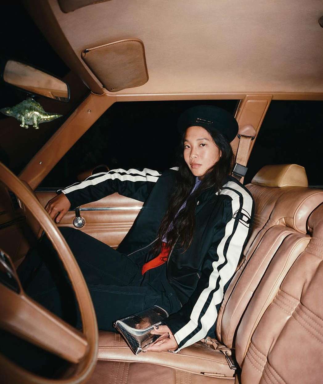 Asian woman in black tracksuit with white stripes and a beret posing in car with vintage brown interior and toy dinosaur.