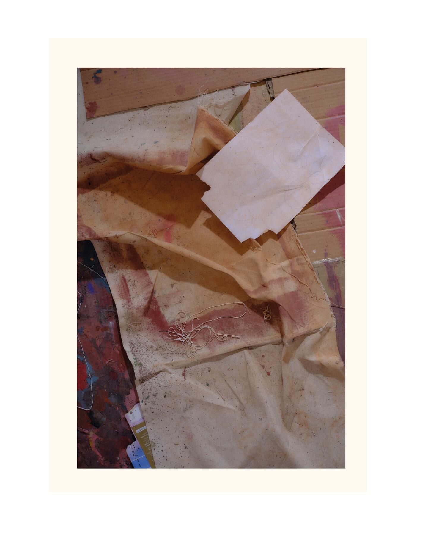 Abstract art piece features folded and distressed beige fabric alongside a small white block on a colorful, paint-splattered surface.