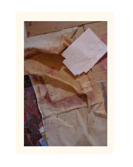 Abstract art piece features folded and distressed beige fabric alongside a small white block on a colorful, paint-splattered surface.