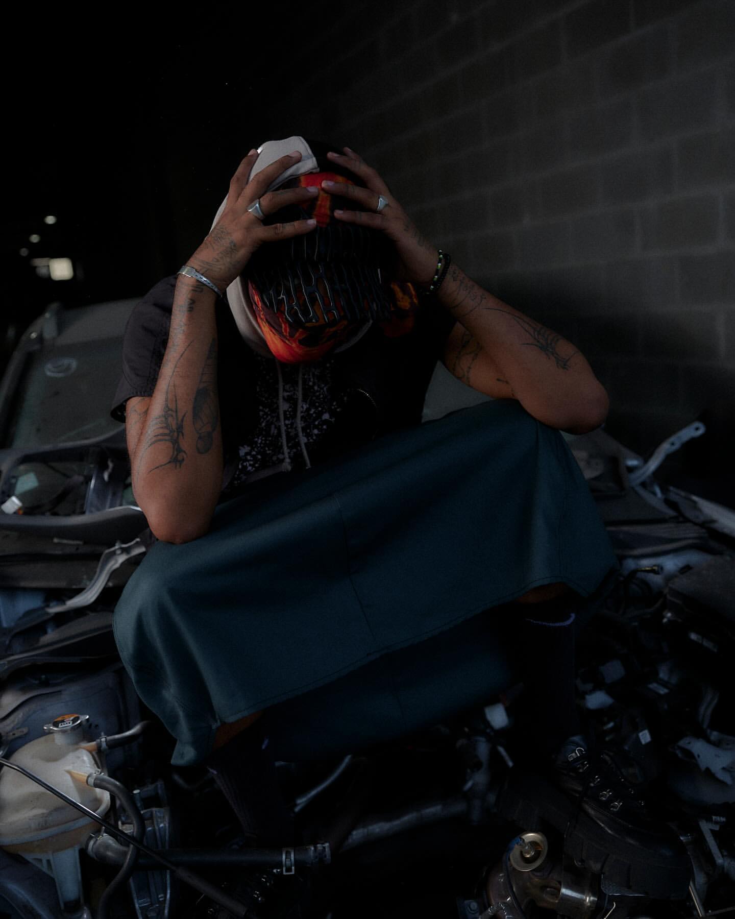 Adult male with tattoos sits hunched on pile of car parts, holding his head, and wearing a helmet, t-shirt, and skirt in an urban environment.