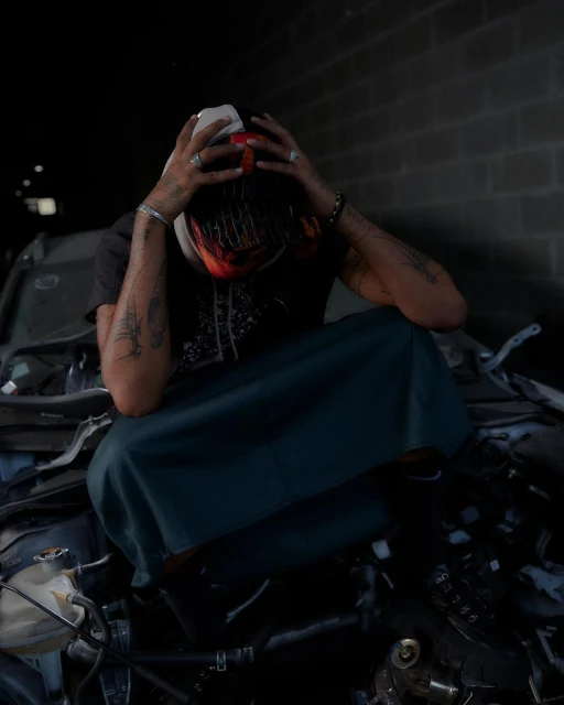 Adult male with tattoos sits hunched on pile of car parts, holding his head, and wearing a helmet, t-shirt, and skirt in an urban environment.
