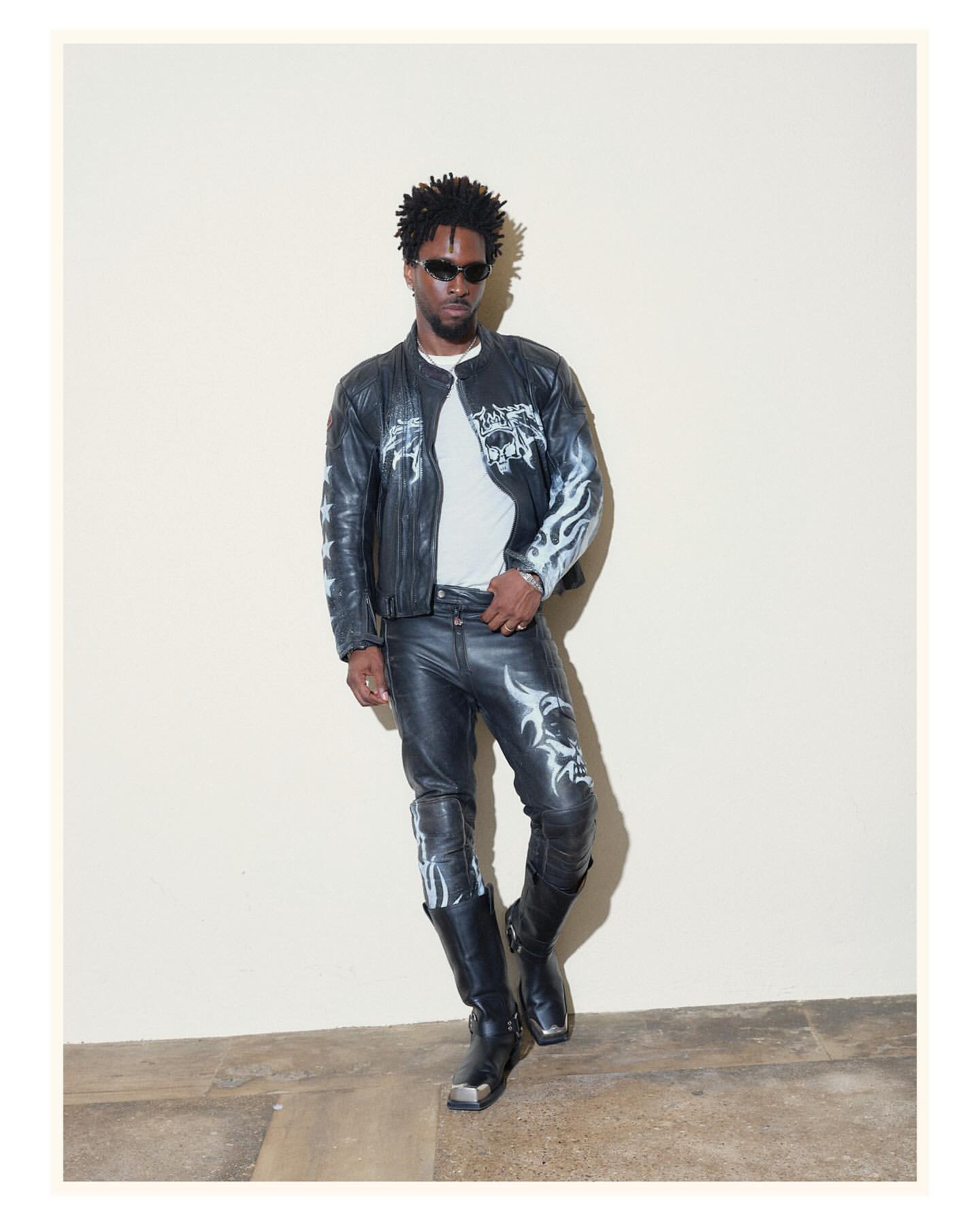 A stylish Black man in a black leather jacket and pants with silver flame designs poses against an off-white wall wearing sunglasses.