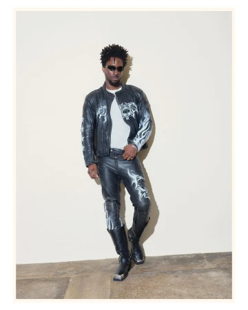 Man wearing a black leather jacket with white graphics, matching leather pants, and boots against a plain wall.