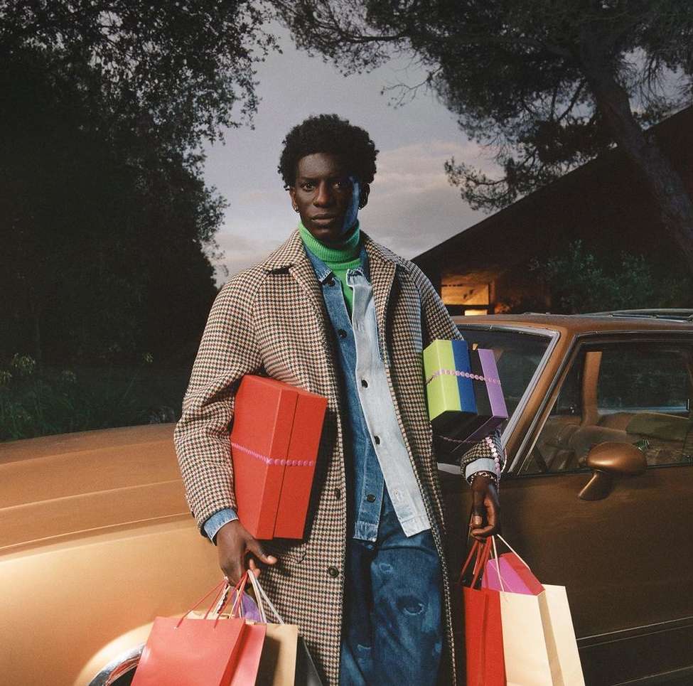 A Black male in a brown plaid coat stands outside by a gold car, holding shopping bags and a box with gift ribbon, in a fashion editorial.