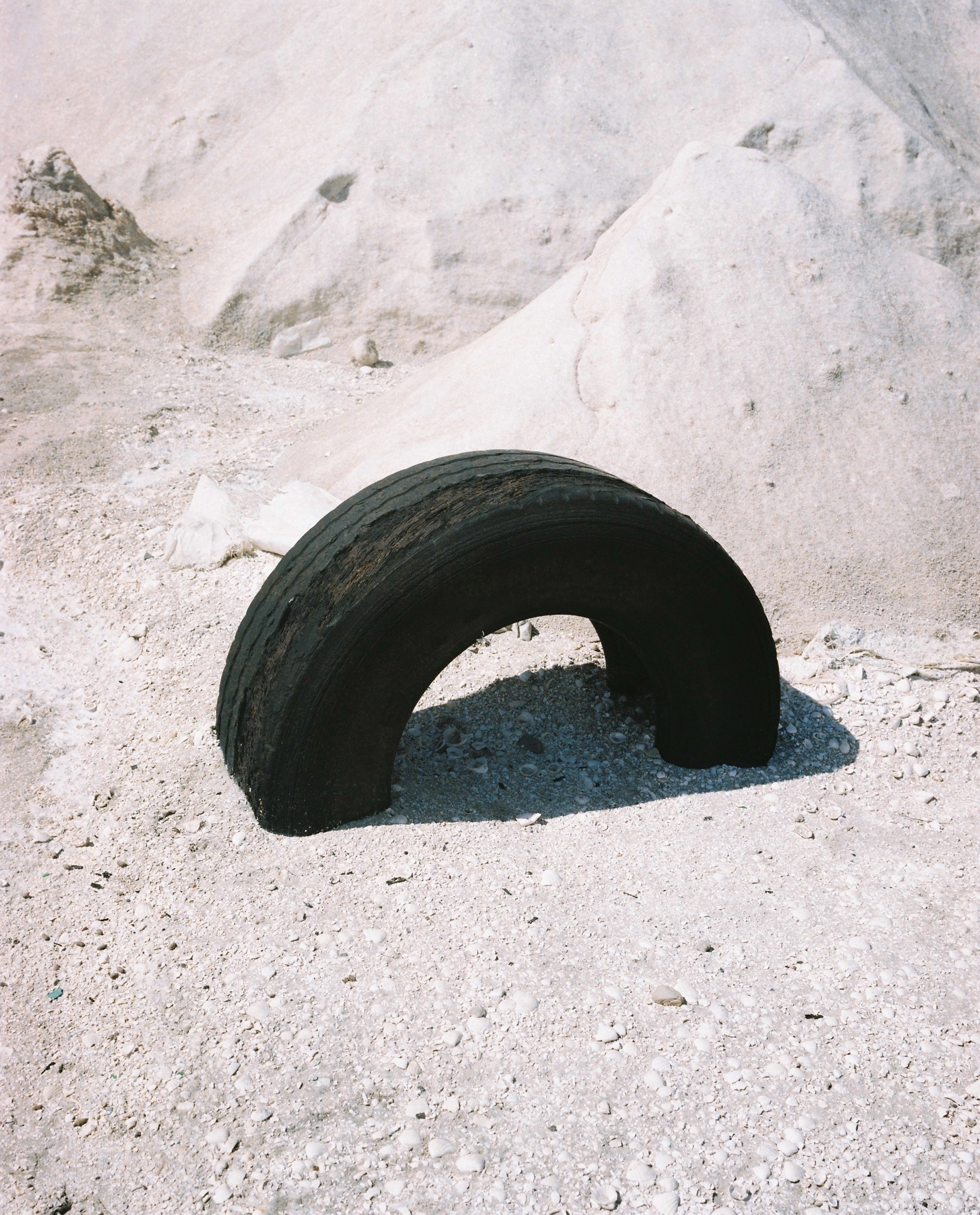 Worn black tire rests on sandy ground against a backdrop of pale dunes, creating a minimalist scene with textures and muted tones.