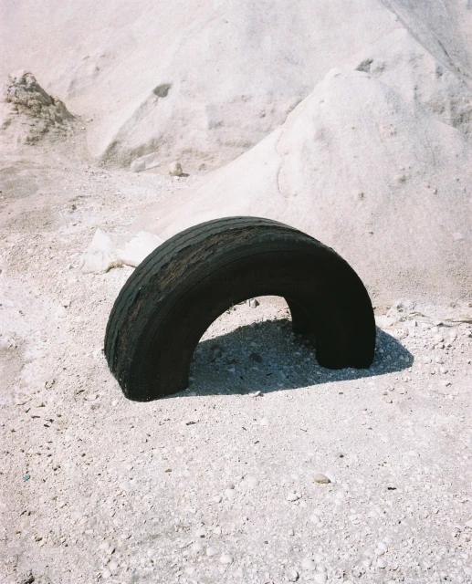 Worn black tire rests on sandy ground against a backdrop of pale dunes, creating a minimalist scene with textures and muted tones.