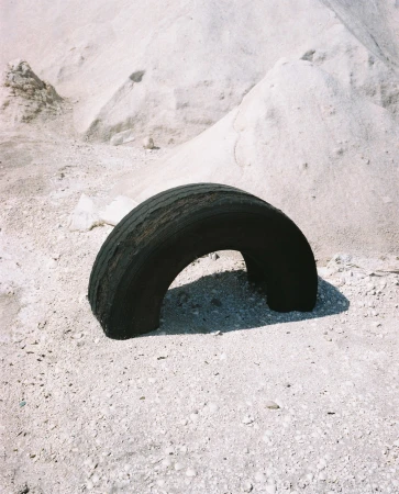 Worn black tire rests on sandy ground against a backdrop of pale dunes, creating a minimalist scene with textures and muted tones.