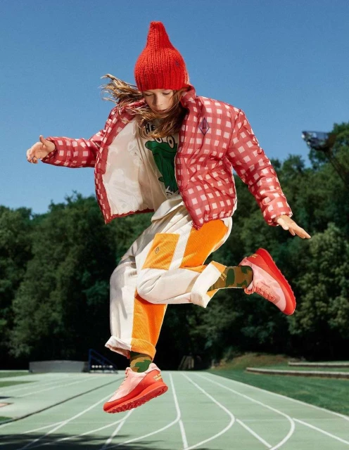 A young person jumping on an outdoor track, wearing a red hat, red and white checkered coat, and matching red shoes, with trees in background.