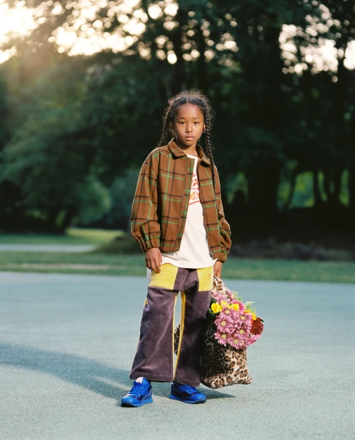 Full-length portrait of an African-descent child in a plaid jacket, wide-leg pants, and blue shoes holding a floral bag, outdoors.