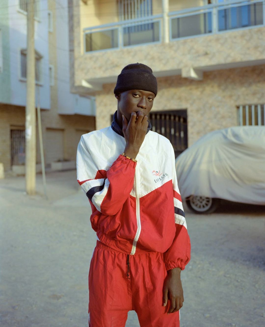 Portrait of an African male in a red and white tracksuit, standing outdoors against an urban background, wearing a black beanie.