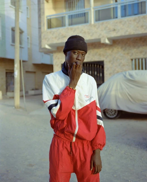 Portrait of an African male in a red and white tracksuit, standing outdoors against an urban background, wearing a black beanie.