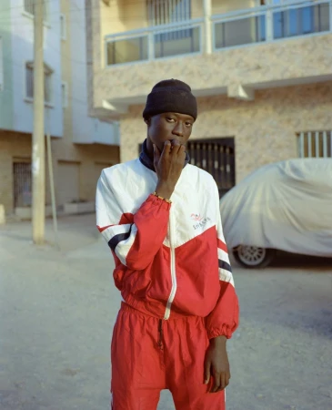 Portrait of an African male in a red and white tracksuit, standing outdoors against an urban background, wearing a black beanie.