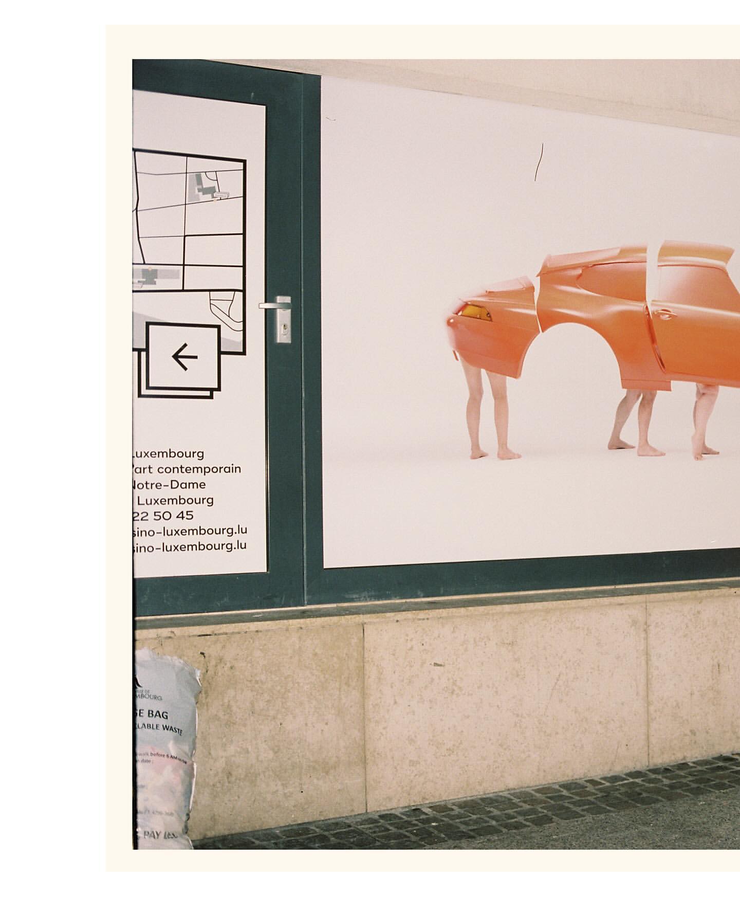 Art installation featuring deconstructed orange car body parts held up by two human legs on a white background, viewed through a window.