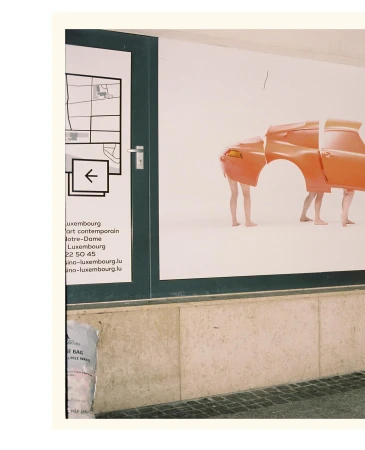 Art installation featuring deconstructed orange car body parts held up by two human legs on a white background, viewed through a window.
