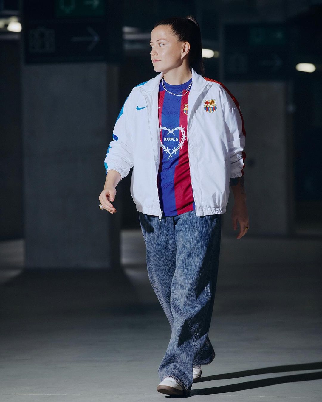 A woman in a white jacket and blue jeans walking in a garage, wearing a soccer jersey. The lighting is soft and diffused.