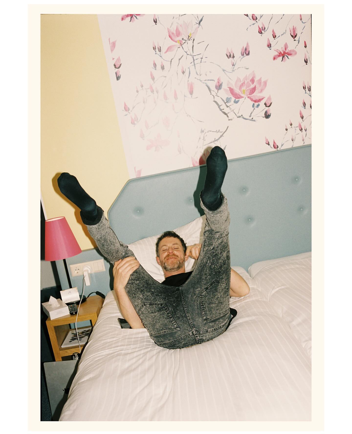 Adult male laying on a bed with his legs in the air, wearing grey jeans and black socks. Pink lamp and floral wallpaper in the bedroom.