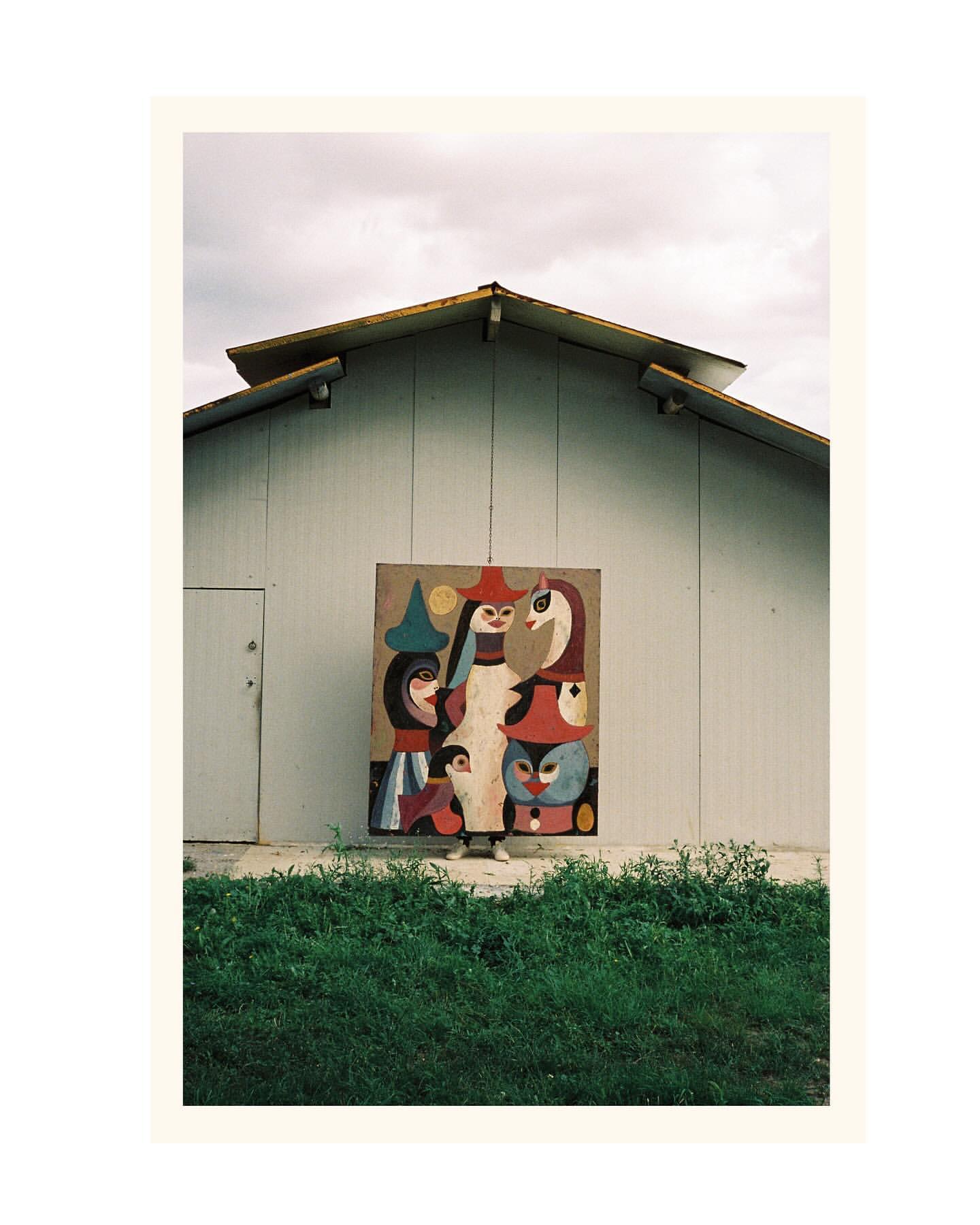 Exterior shot of an abstract painting on a wall of a white building, with a green grassy area in the foreground under a cloudy sky.