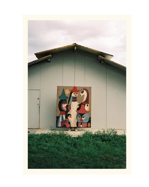 Exterior shot of an abstract painting on a wall of a white building, with a green grassy area in the foreground under a cloudy sky.