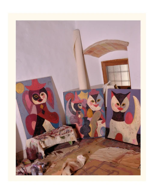 Three colorful paintings of cartoon-like cats and a harlequin character are displayed in an artist's studio, illuminated by natural light.