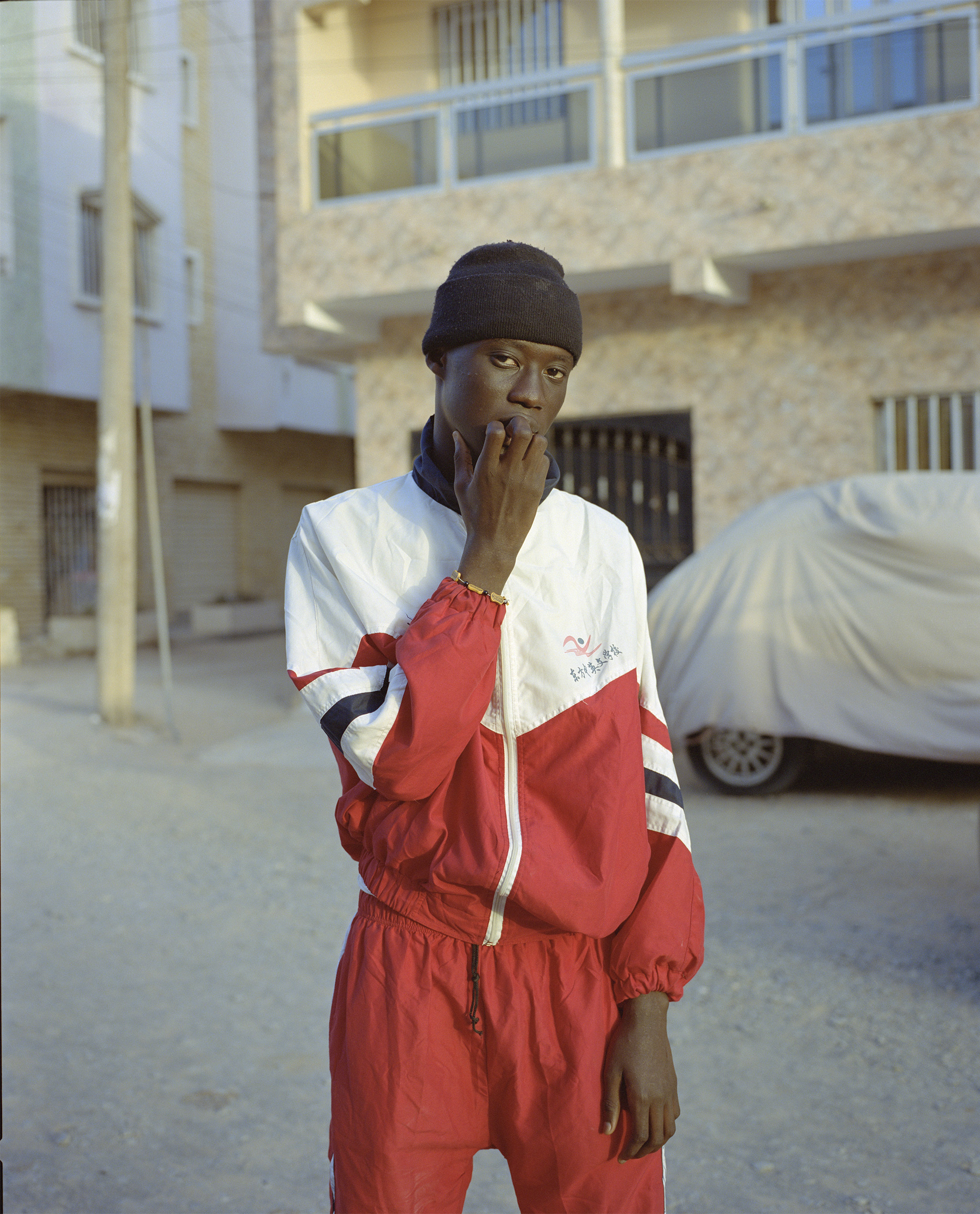 Young adult male in red tracksuit and black beanie stands outside building with beige facade and covered car in an eye-level shot.