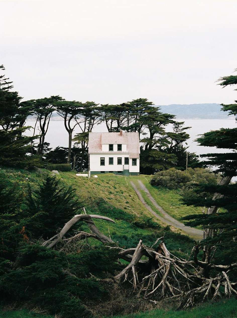 White house with a red roof sits on a green hillside with trees, overlooking a body of water under an overcast sky.