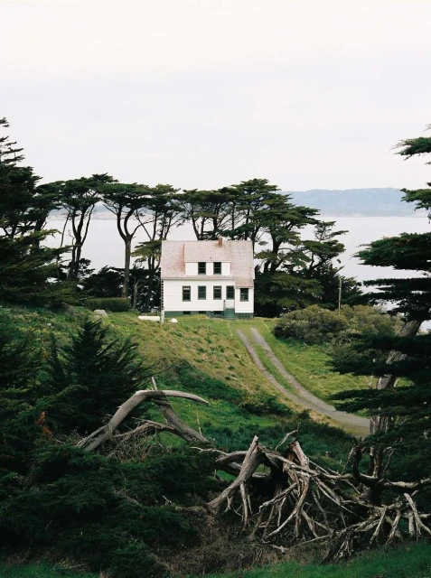 White house with a red roof sits on a green hillside with trees, overlooking a body of water under an overcast sky.