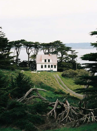 White house with a red roof sits on a green hillside with trees, overlooking a body of water under an overcast sky.