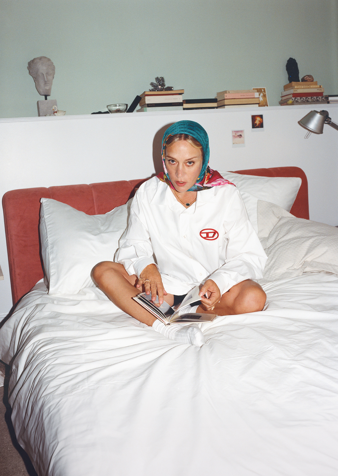 Young woman sits cross-legged on a white bed, headscarf, Diesel branded long-sleeve shirt reading a book in an indoor bedroom setting.