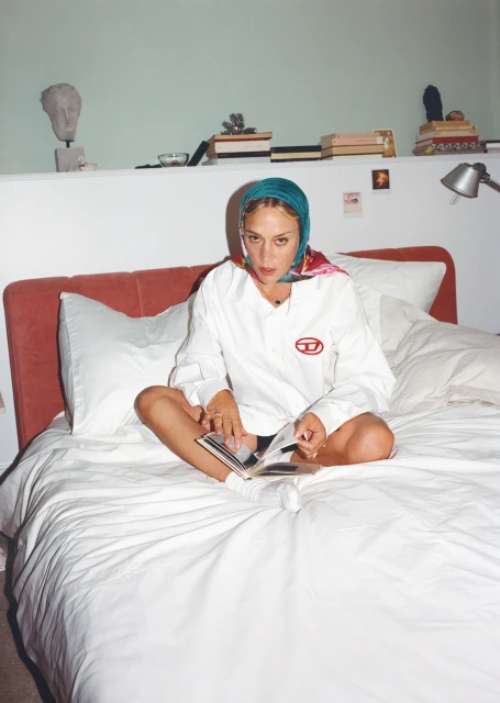 Young woman sits cross-legged on a white bed, headscarf, Diesel branded long-sleeve shirt reading a book in an indoor bedroom setting.