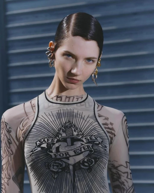 Close-up of a fair-skinned woman with slicked-back dark hair and unique earrings, wearing a sheer top with a tattoo design.