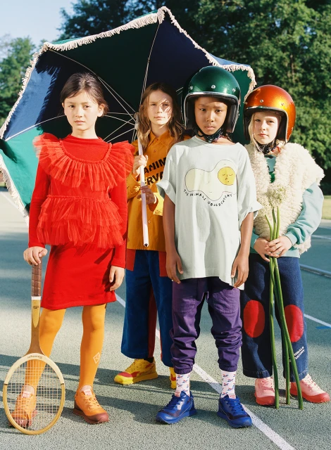 Four children stand outside on a painted surface wearing colorful vintage outfits, helmets, and holding a tennis racket and flowers.