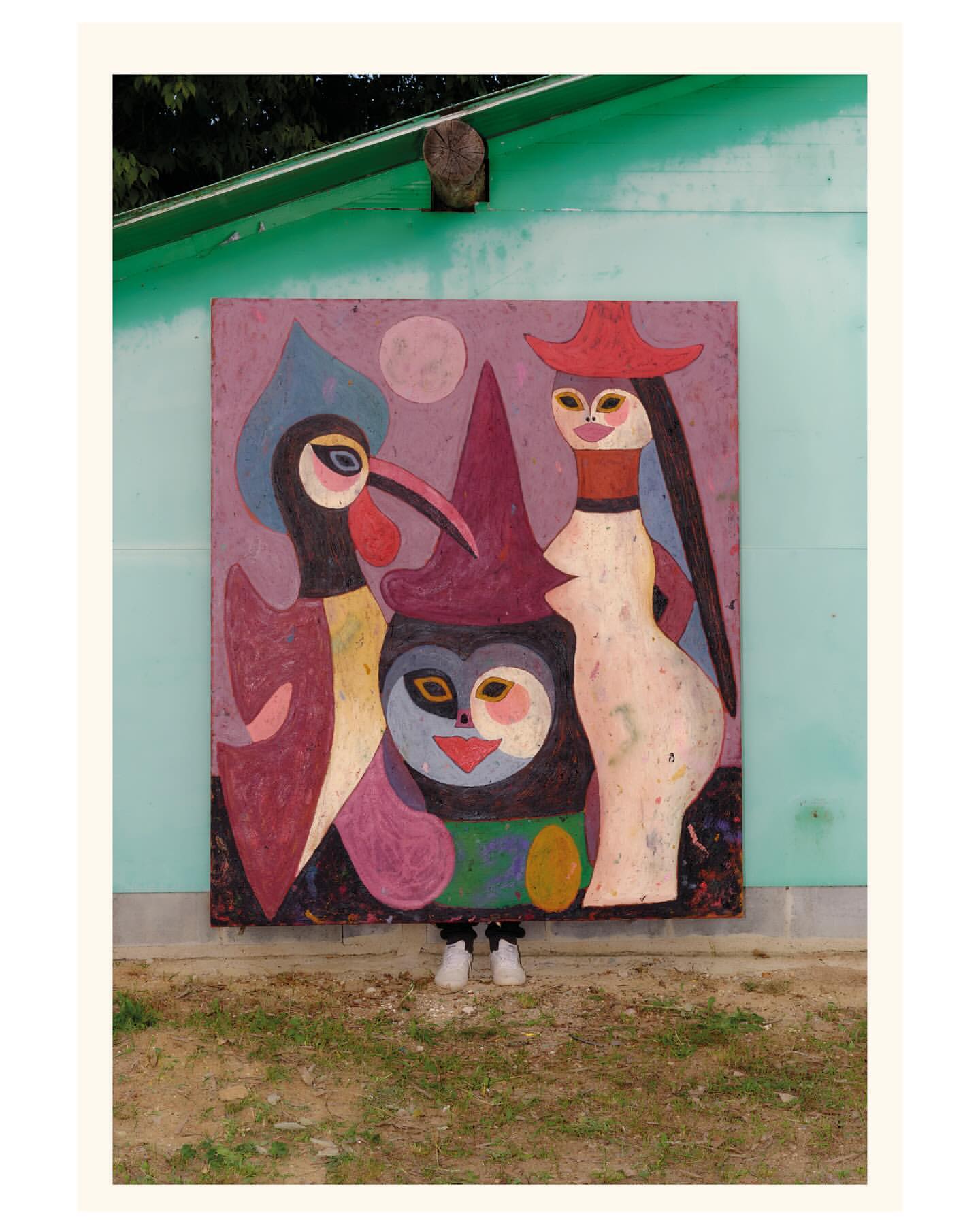 A person hidden behind a large painted artwork featuring stylized figures mounted on a teal wall outdoors