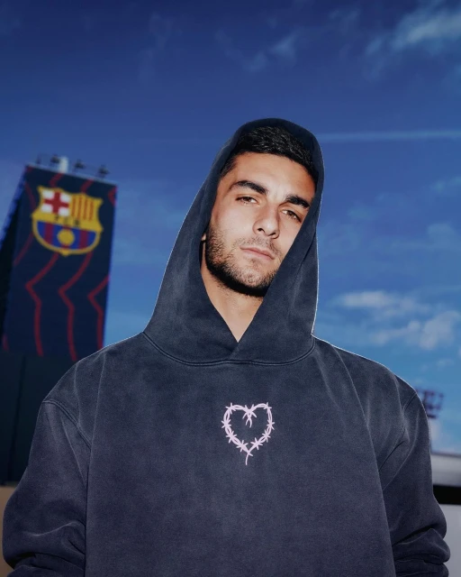Portrait of a young adult male in a hooded sweatshirt with a barbed wire heart design, standing outdoors with a soccer stadium.