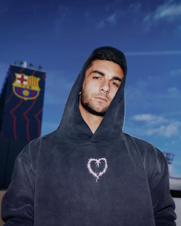 Portrait of a young adult male in a hooded sweatshirt with a barbed wire heart design, standing outdoors with a soccer stadium.
