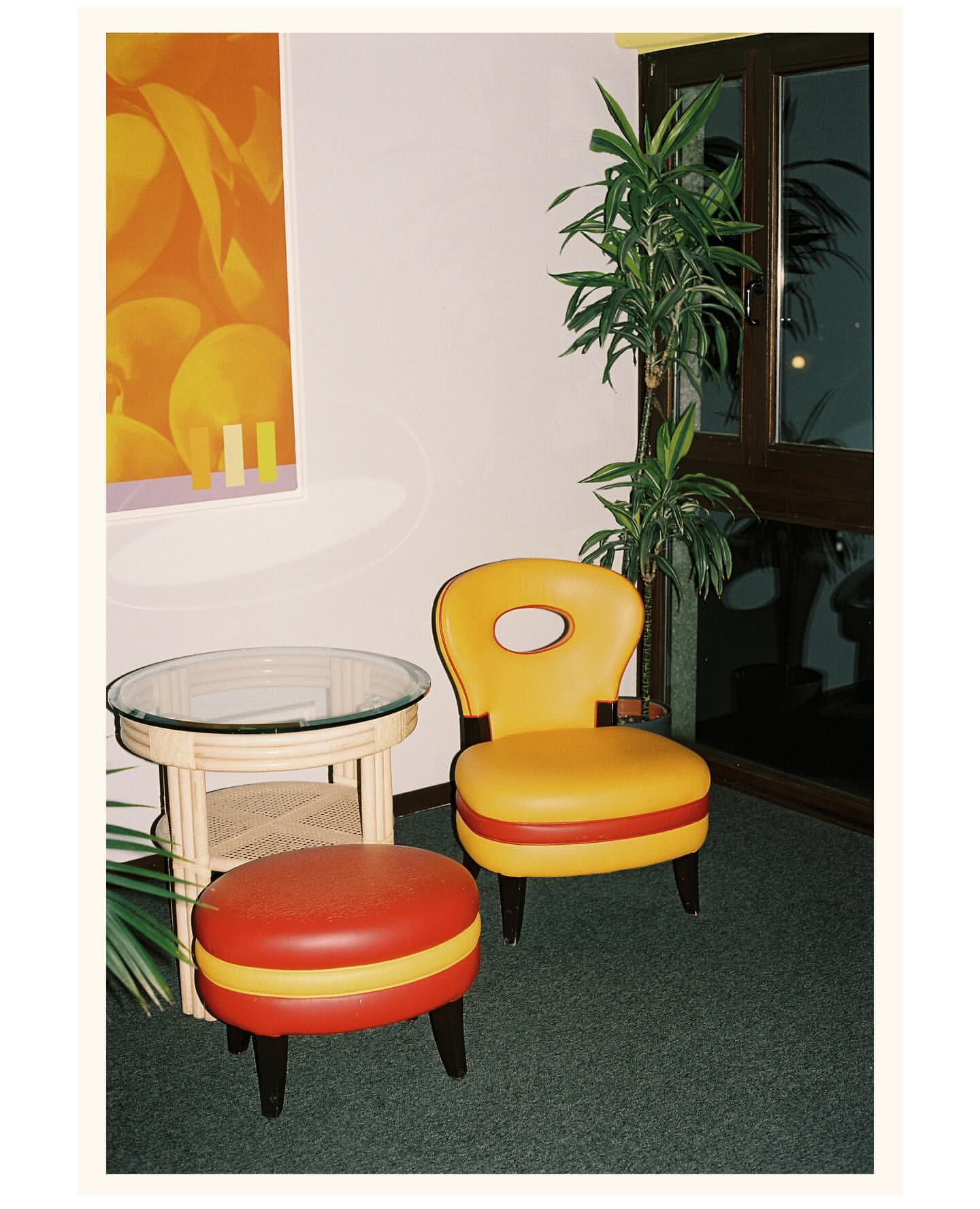 Retro furniture set in a corner featuring a yellow chair, red and yellow ottoman, and a glass-topped table, complemented by an orange wall art.