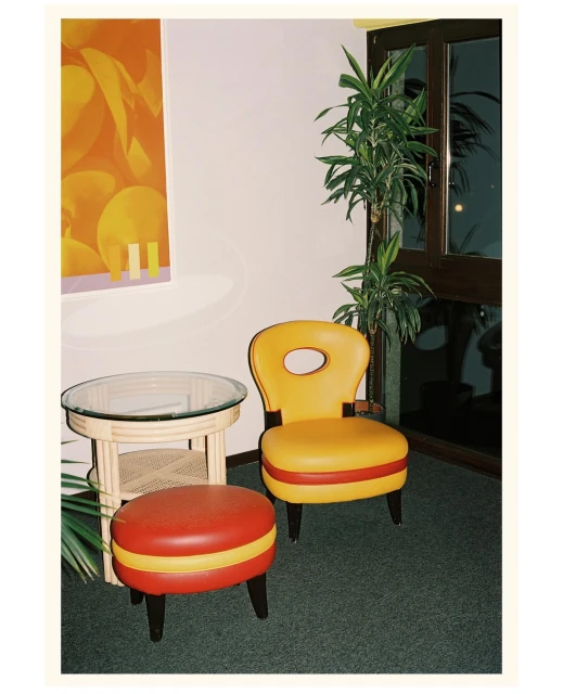 Retro furniture set in a corner featuring a yellow chair, red and yellow ottoman, and a glass-topped table, complemented by an orange wall art.