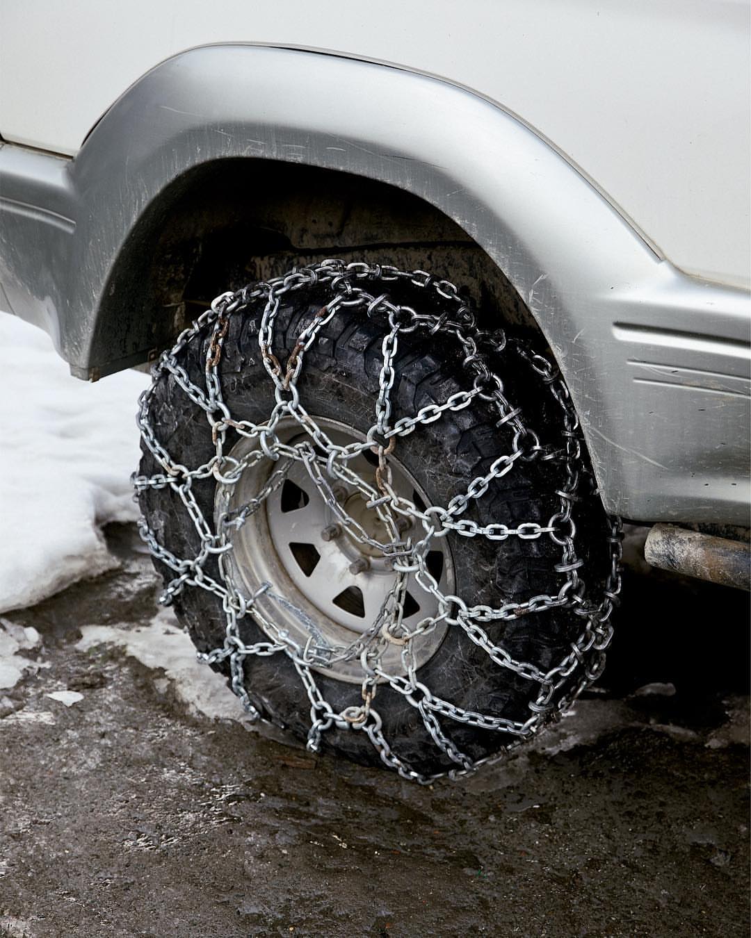 Close-up of a vehicle's tire fitted with snow chains, providing enhanced traction on an icy surface. The gray car is partially visible.