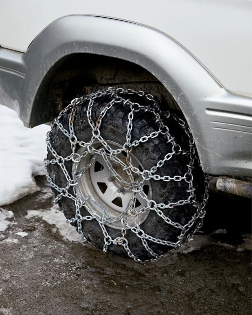 Close-up of a vehicle's tire fitted with snow chains, providing enhanced traction on an icy surface. The gray car is partially visible.