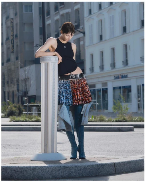 Woman in black tank top and pleated skirt leaning on a bollard, with city buildings visible in the background, in an outdoor setting.