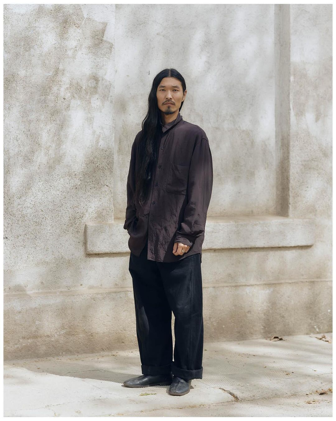 Portrait of an adult Asian male with long hair wearing a brown button-down shirt and dark trousers standing outdoors against a concrete wall.