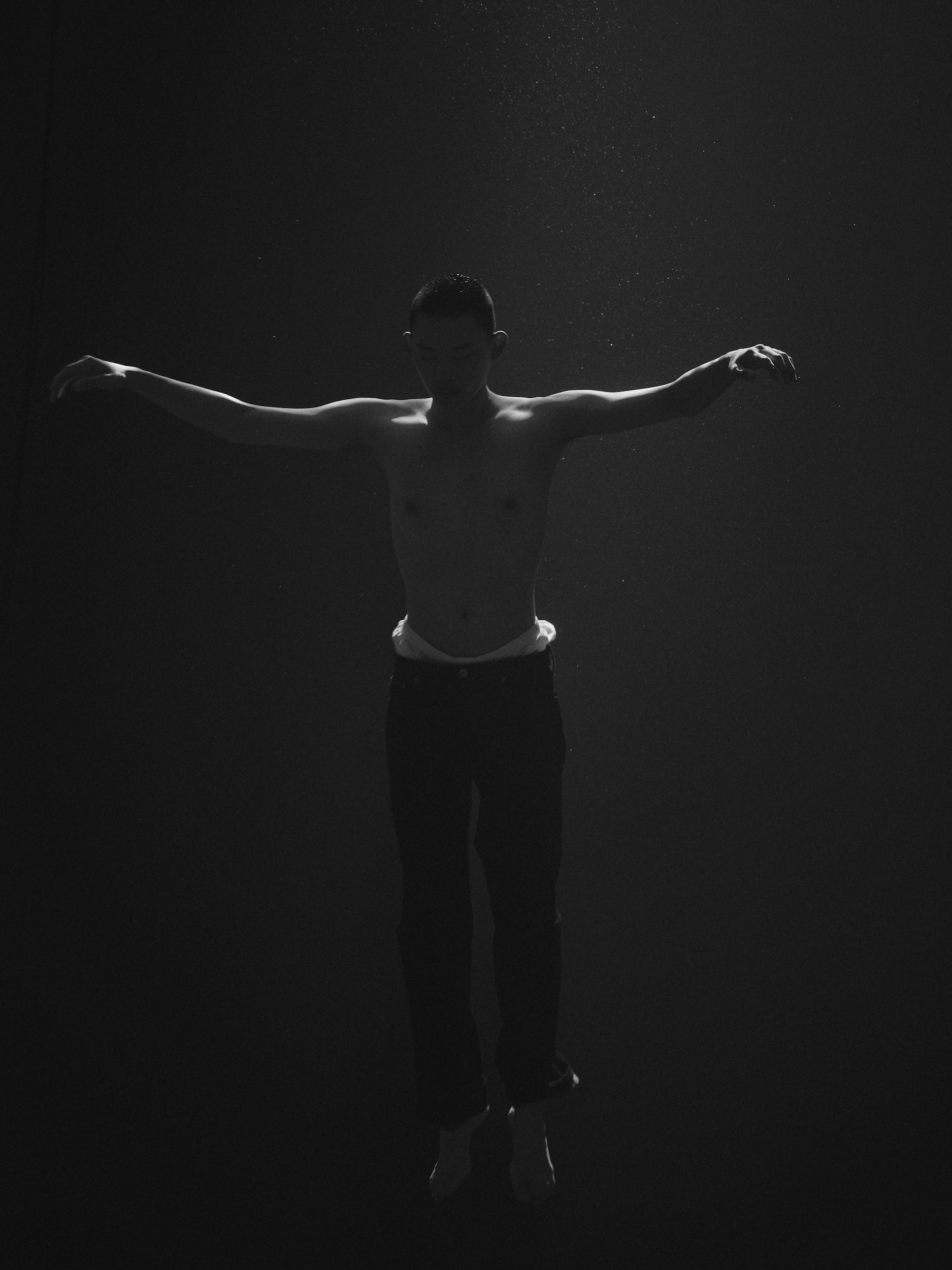 Monochrome underwater portrait of a shirtless man floating with arms outstretched in dark murky water by Dani Pujalte.