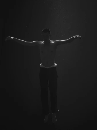Monochrome underwater portrait of a shirtless man floating with arms outstretched in dark murky water by Dani Pujalte.