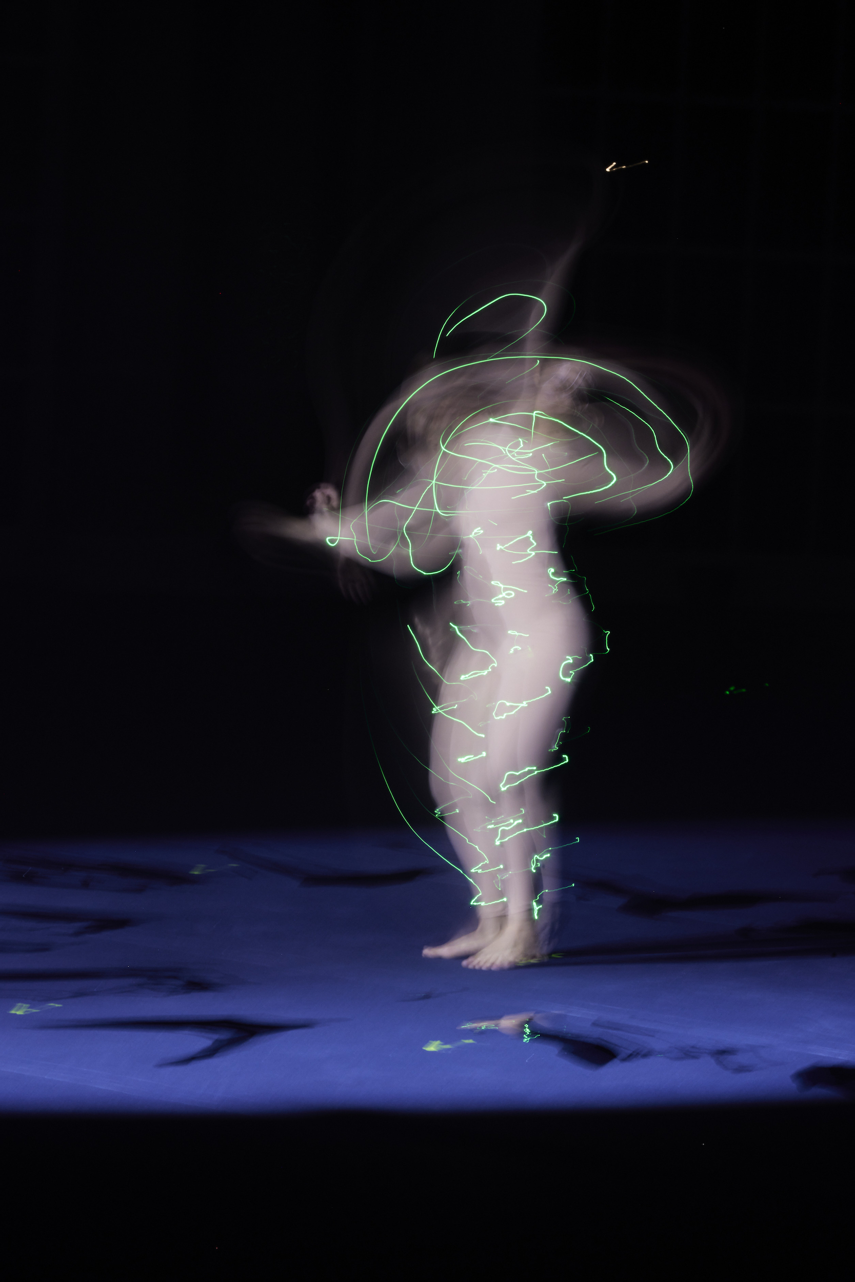 Solo adult female stands on a blue surface in the dark, body encircled with neon-green light trails from long-exposure photography.
