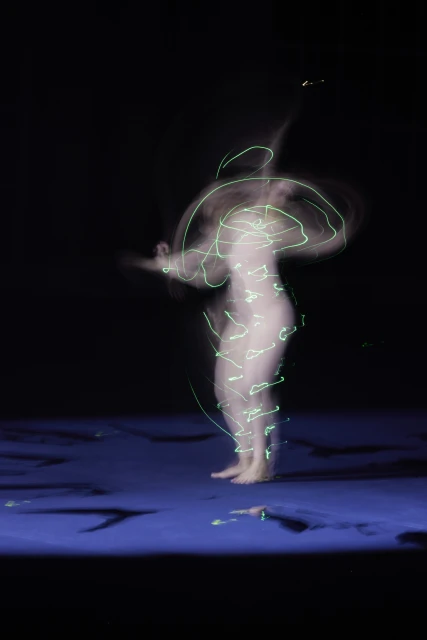 Solo adult female stands on a blue surface in the dark, body encircled with neon-green light trails from long-exposure photography.