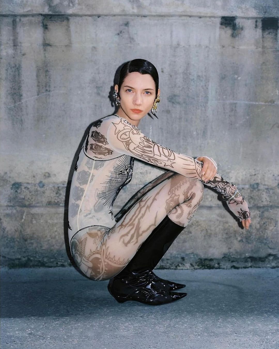 A young woman with sleek hair poses in a printed bodysuit and black patent boots against a textured gray wall in an outdoor setting.