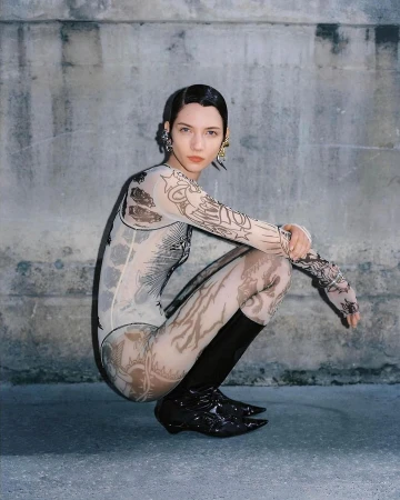 A young woman with sleek hair poses in a printed bodysuit and black patent boots against a textured gray wall in an outdoor setting.